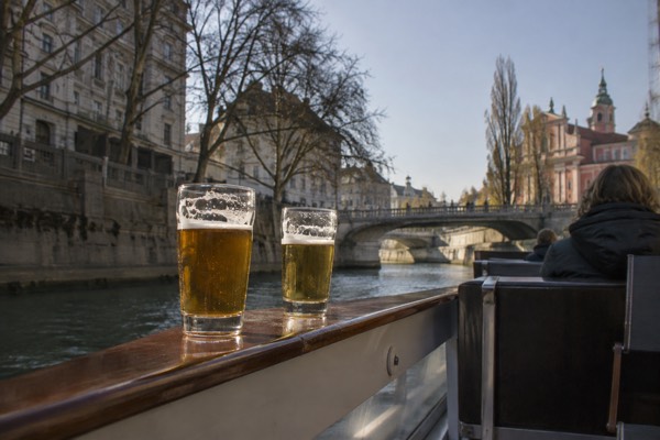 Glasses of craft beer on the rail of an open boat moving along a river between stone embankments, Ljubljana City Centre, Ljubljana