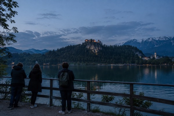 A small group at a lakeside railing looking across still green water to wooded slopes and a distant hilltop, Lake Bled, Bled