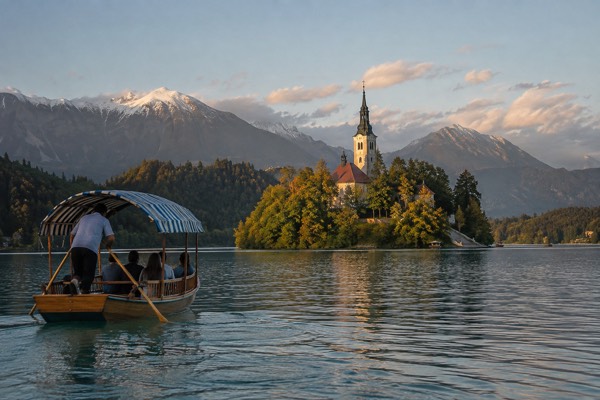A wooden boat with a striped awning being rowed across a green lake towards a small wooded island, Lake Bled, Bled