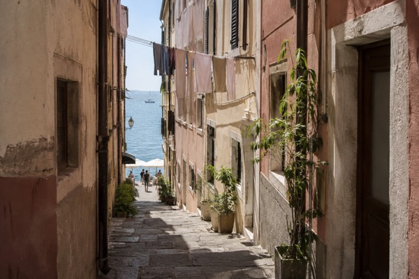 A tight lane of tall pastel houses running down to a sunlit sea front, laundry lines overhead and open water at the end, Piran, Slovenia