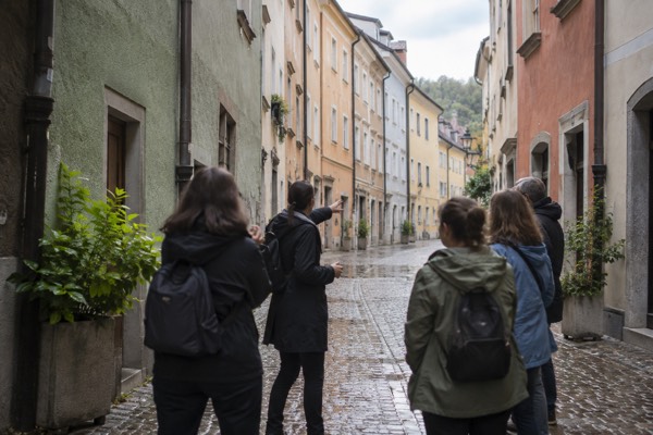 A guide pointing along a narrow street of tall painted house fronts while a small group listens, Poljane, Ljubljana