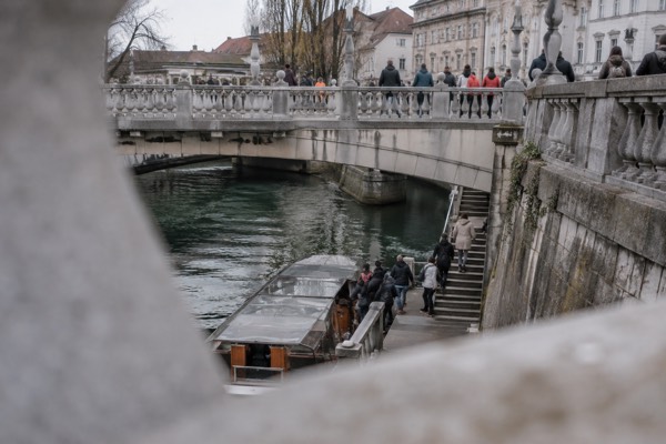 A group stepping off a low boat onto stone river steps beneath linked footbridges, their guide waiting above, Triple Bridge, Ljubljana