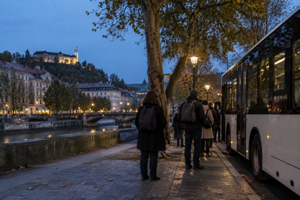 People waiting with day bags at a stop under plane trees, the plain side panel of a city bus filling one edge of the frame, Ljubljana City Centre, Ljubljana