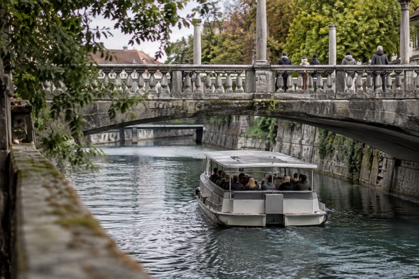 A low open electric boat gliding along a river between stone embankments, passengers looking up as it slips under a footbridge, Cobblers' Bridge, Ljubljana