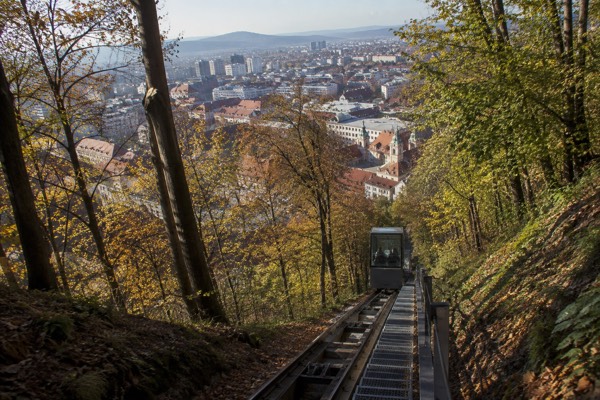 A glazed funicular carriage climbing a steep wooded track towards the top of a castle hill, the city dropping away behind it, Ljubljana Castle Funicular, Ljubljana