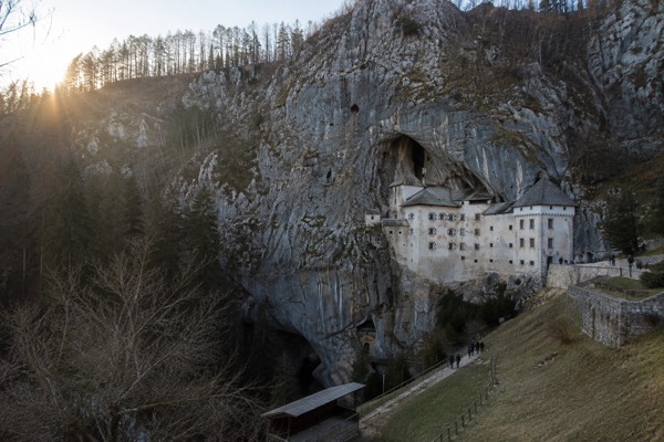 A stone castle front set into the mouth of a pale limestone cliff, bare trees at its foot and visitors small below it, Predjama Castle, Predjama
