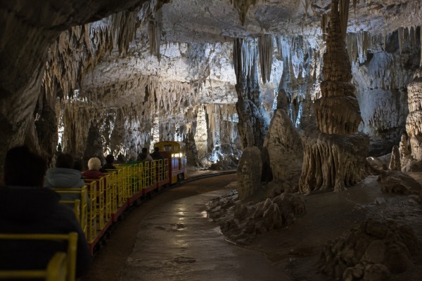 A small visitor train running into a lit limestone chamber deep underground, passengers seated in open carriages, Postojna Cave, Postojna