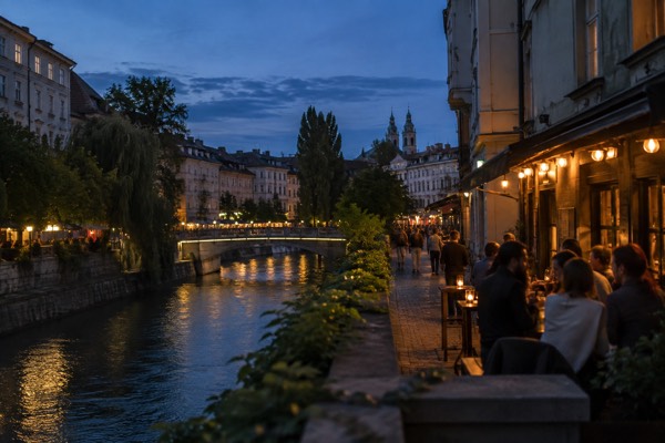 A group with drinks outside a bar on a narrow city street after dark, warm light spilling across the pavement, Ljubljana City Centre, Ljubljana