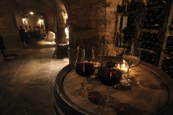 Glasses of red and white wine on a barrel top in a low vaulted brick cellar, bottles racked in the dark behind, Ljubljana Old Town, Ljubljana
