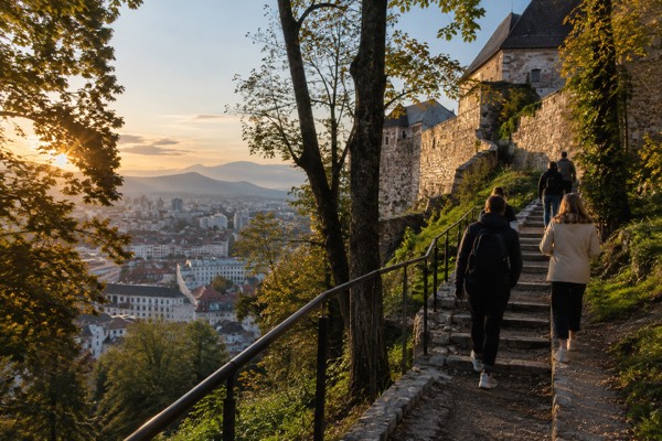 A group climbing a stepped woodland path towards castle walls, the city rooftops falling away behind them, Ljubljana Castle, Ljubljana