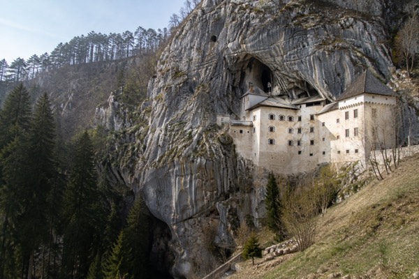 A castle wall built flush into a pale limestone overhang, scree and bare trees below it, Predjama Castle, Predjama