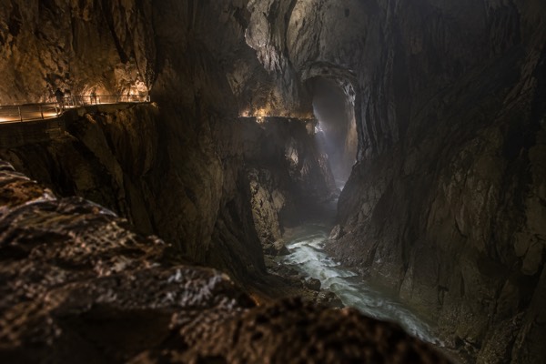 A narrow walkway cut into the wall of a vast underground limestone chamber, tiny figures on it far off, Skocjan Caves, Divaca