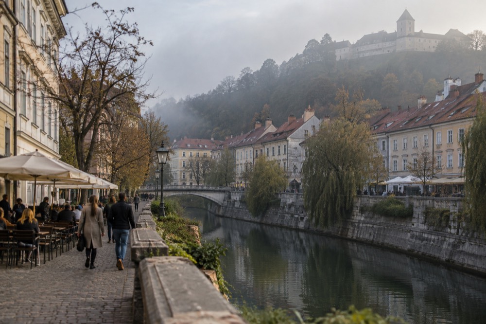 Riverside buildings along the Ljubljanica with cafe terraces and the Ljubljana castle hill rising behind