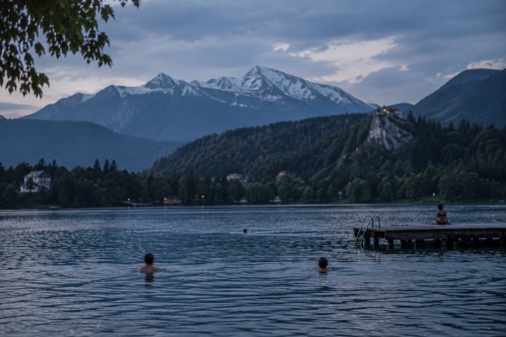 Swimmers in a calm Alpine lake near a wooden platform with green forest around the shore