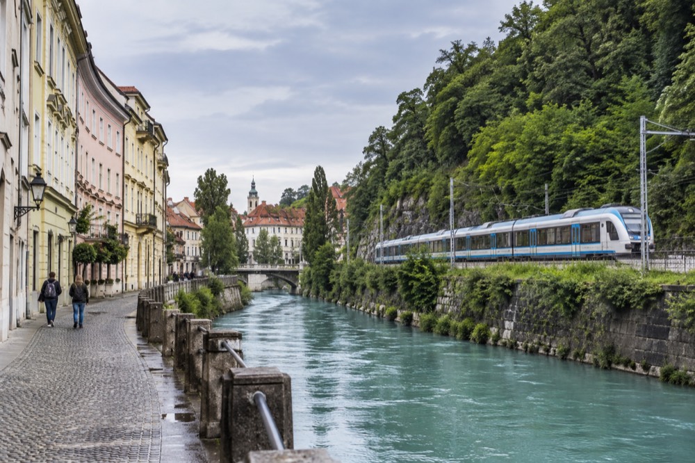 A passenger train travelling along a rail line beside a turquoise river between steep forested slopes