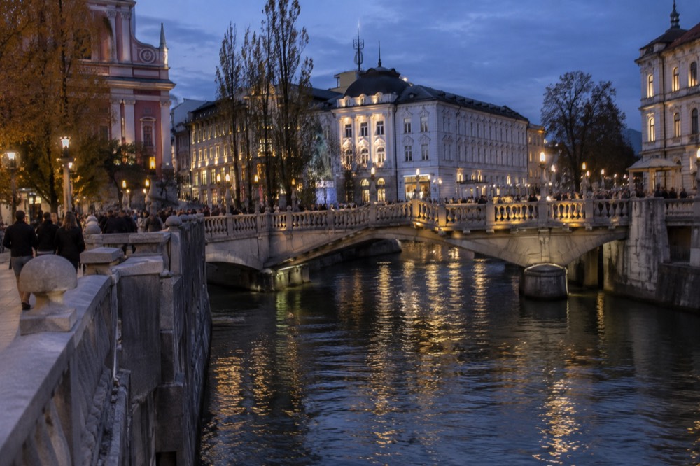 The Ljubljanica river and the Triple Bridge seen from the bank at dusk, with lit facades behind