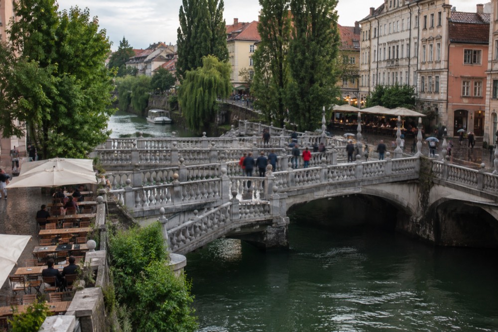 Tables and chairs beside the Ljubljanica river in Ljubljana, with the triple bridge and old buildings along the bank