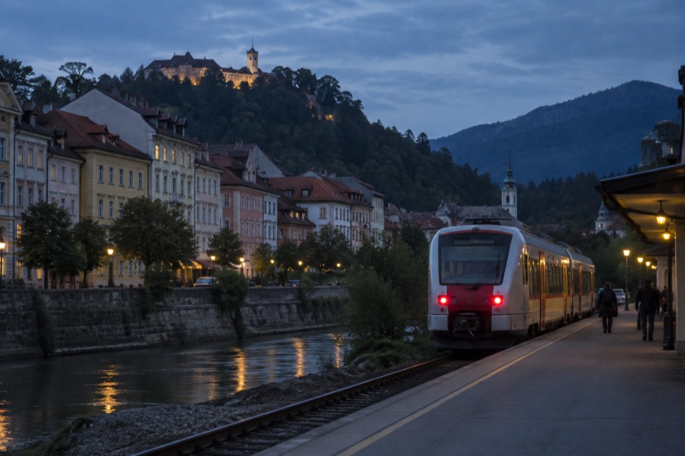 A two-car local train waiting at a small alpine station platform with wooded mountains rising behind it