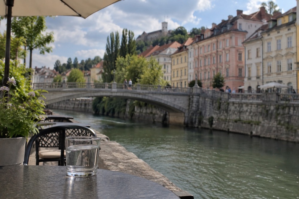 A glass of water on a cafe table beside a river with a stone bridge in the background