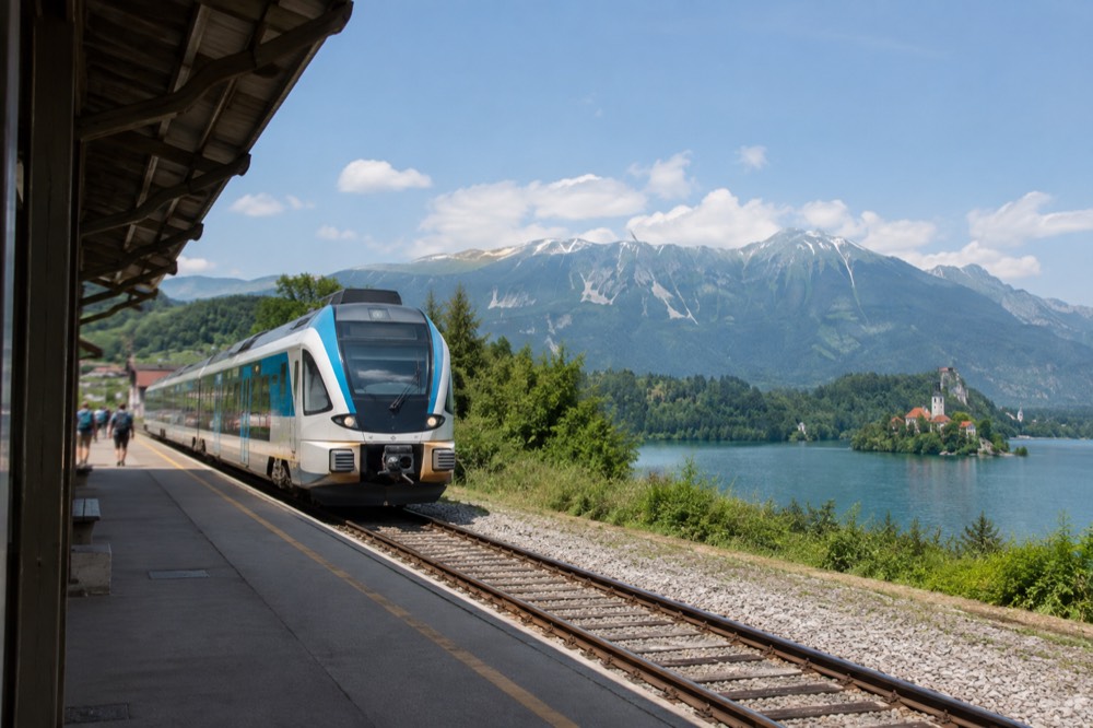 A Slovenian passenger train at a rural station with green hills behind it