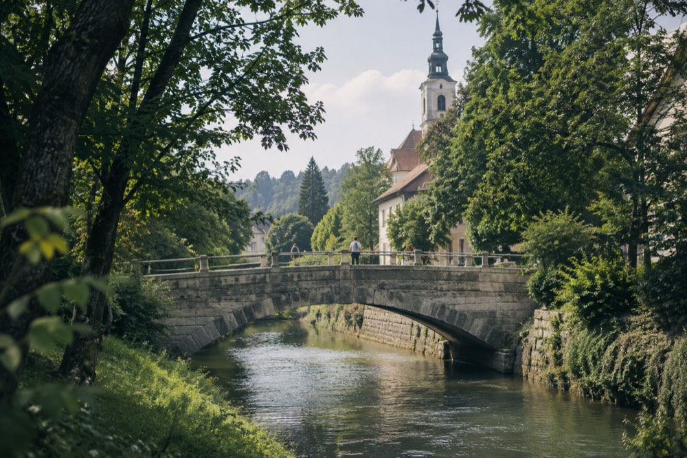 Stone bridge over a small stream with trees on both banks and a church tower beyond