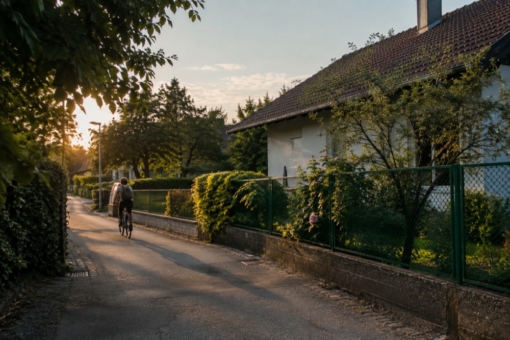 Cyclist passing a low suburban house with a green garden fence on a quiet street