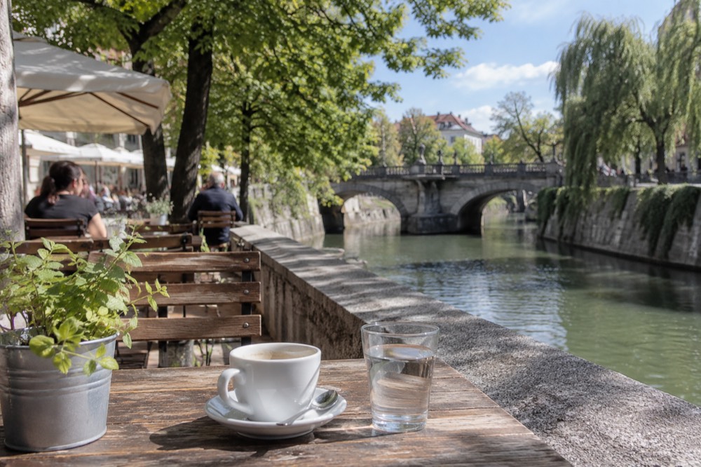 Outdoor café tables along a riverside embankment with trees and an old stone bridge in the background