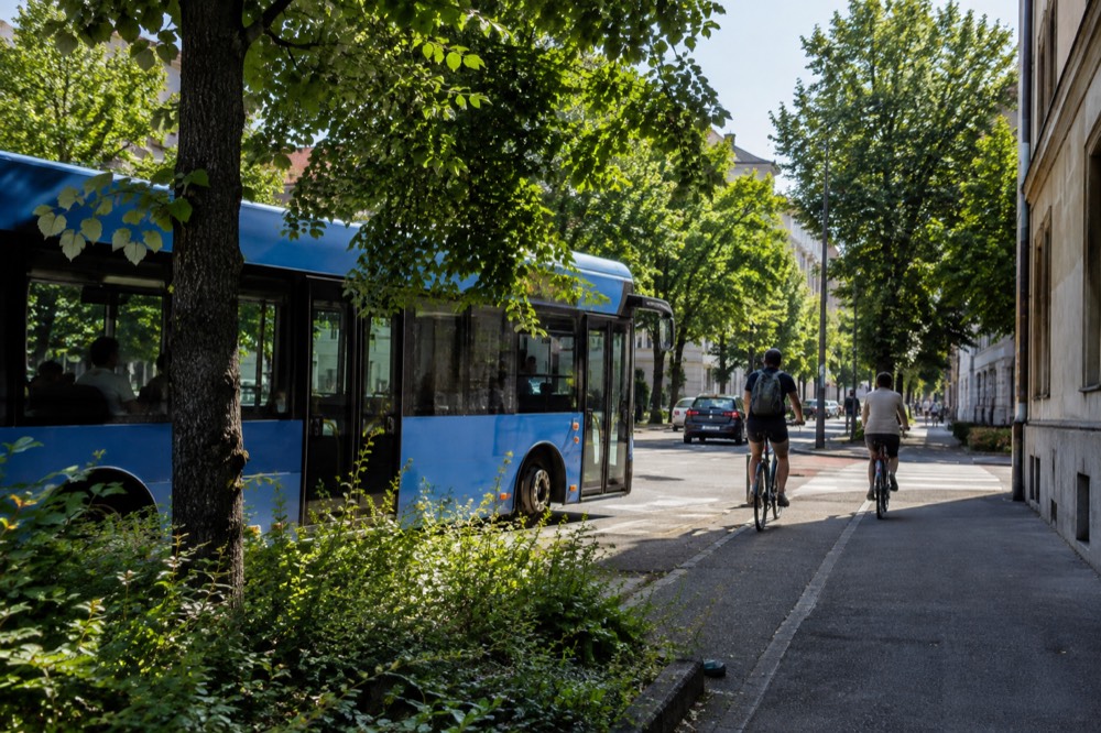Blue city bus stopping at a leafy street corner with cyclists waiting beside it