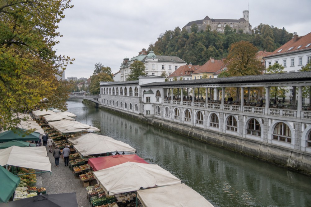 A riverfront arcade with stone columns beside a calm river and a hilltop castle in the background