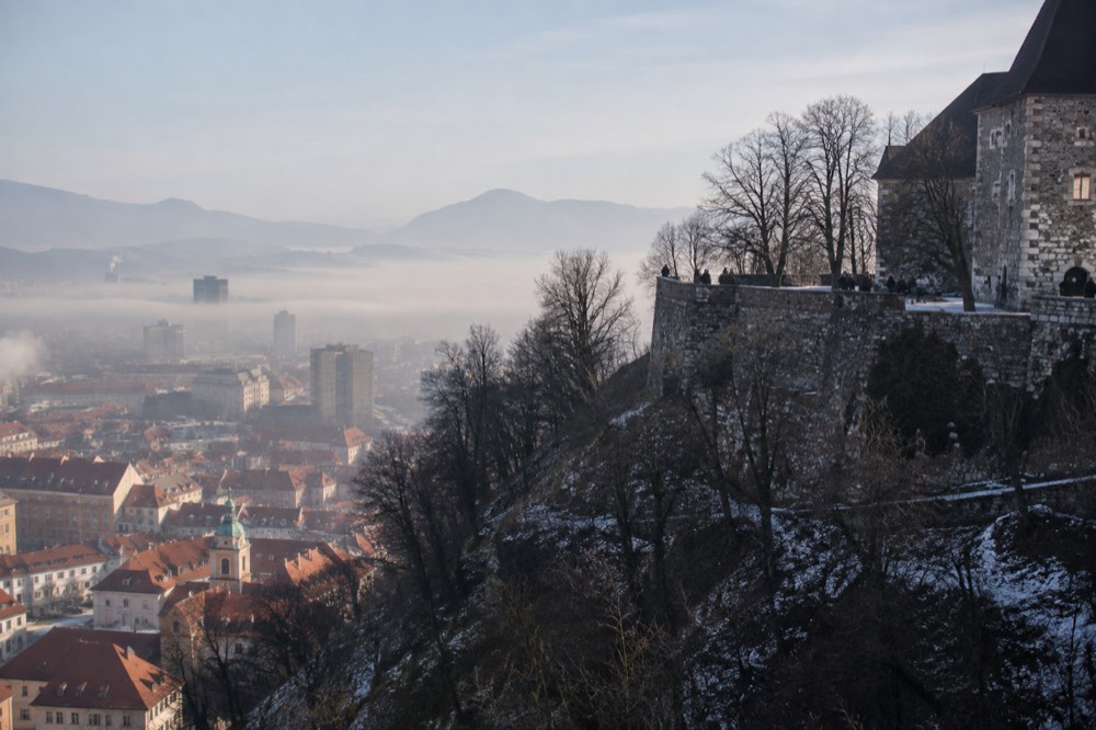 A city skyline of red roofs under low morning fog with a hilltop castle above the mist