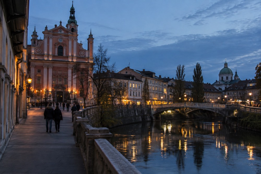 A river at dusk with a domed church silhouette and lit bridge lamps along the bank