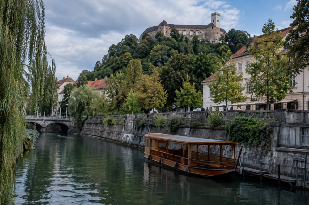 A wooden river boat moored on the Ljubljanica beside a stone embankment with the Ljubljana castle hill rising behind