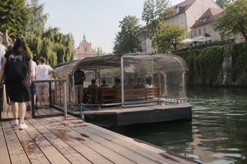 A covered river cruise boat moored at a wooden pier on the Ljubljanica with passengers boarding