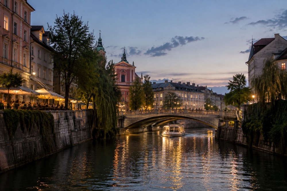 Golden evening light on the Ljubljanica river with a cruise boat passing under a stone bridge and lit facades along the bank
