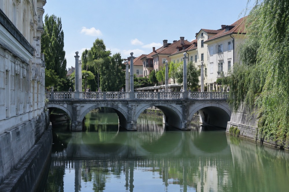 A stone bridge with three parallel spans crossing a green river between pastel buildings