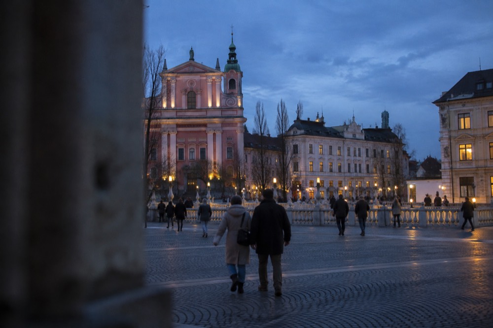Open pedestrian square in central Ljubljana with a church facade and a stone bridge over a river