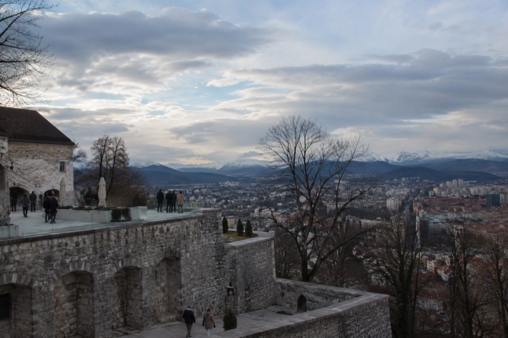 A view from a hilltop terrace across a city with a castle hill and distant snowy mountains