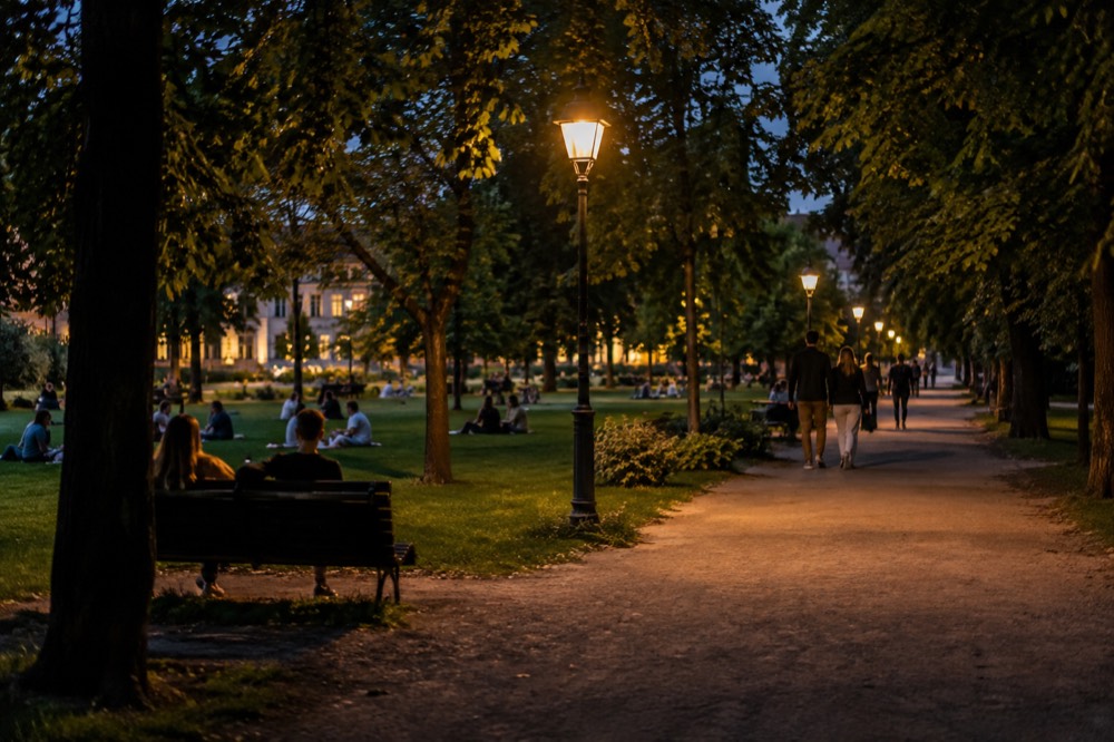 People strolling and sitting on lawns beside a broad park path in evening light