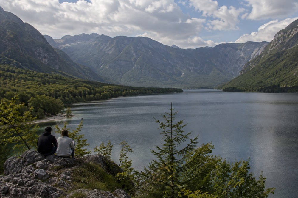 A calm mountain lake at midday with wooded slopes and grey limestone peaks above