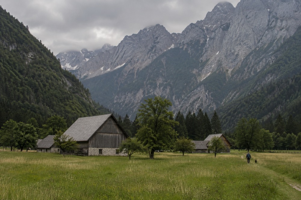 Green meadows on a valley floor with traditional wooden farm buildings and grey limestone cliffs rising behind them