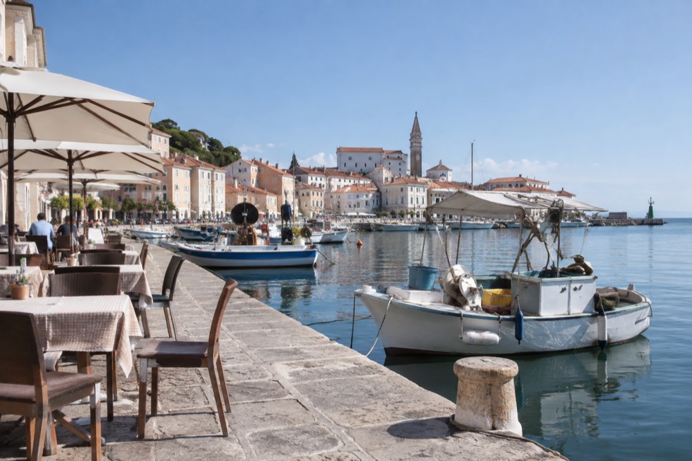 Outdoor restaurant tables beside fishing boats along a stone harbour in Piran
