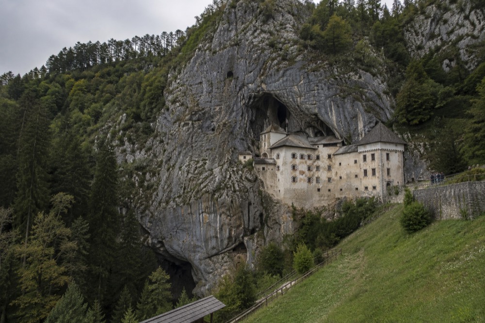 A stone castle facade set into a tall grey limestone cliff with wooded slopes below