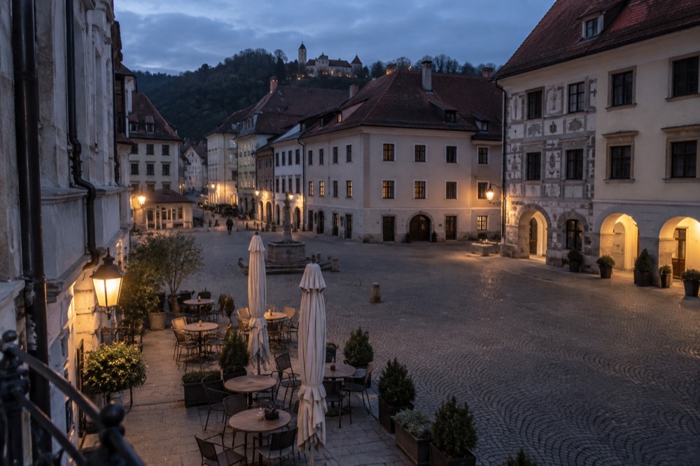 An empty cobbled square with outdoor cafe tables and medieval facades in late afternoon light