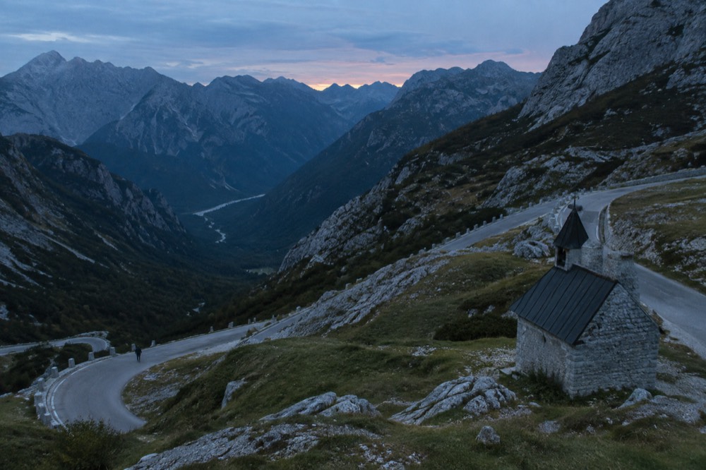 A high mountain pass road curving between grey peaks with a small stone chapel visible
