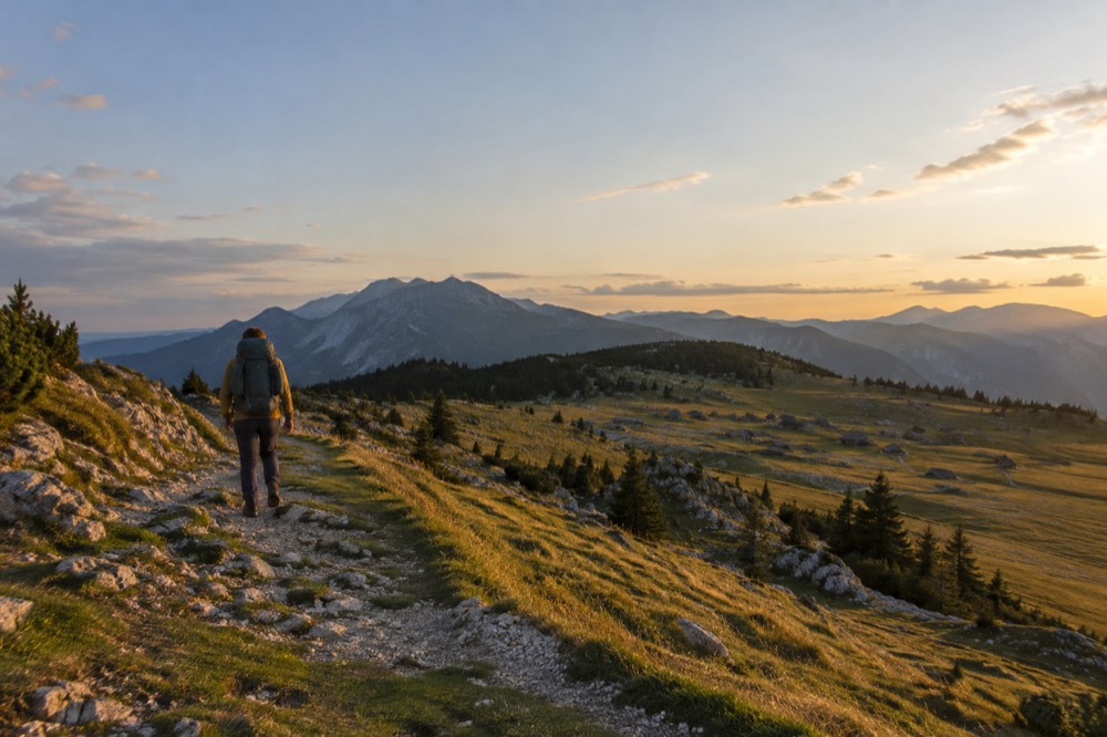 A hiker with a backpack walking a ridge path above a wide alpine meadow