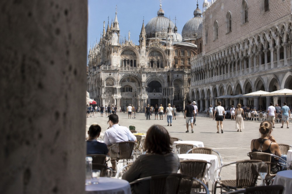 A wide stone square with a basilica facade and café tables under arcades