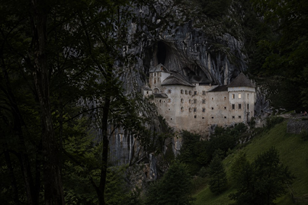 A stone castle built into the mouth of a cliff-face cave above a forested valley