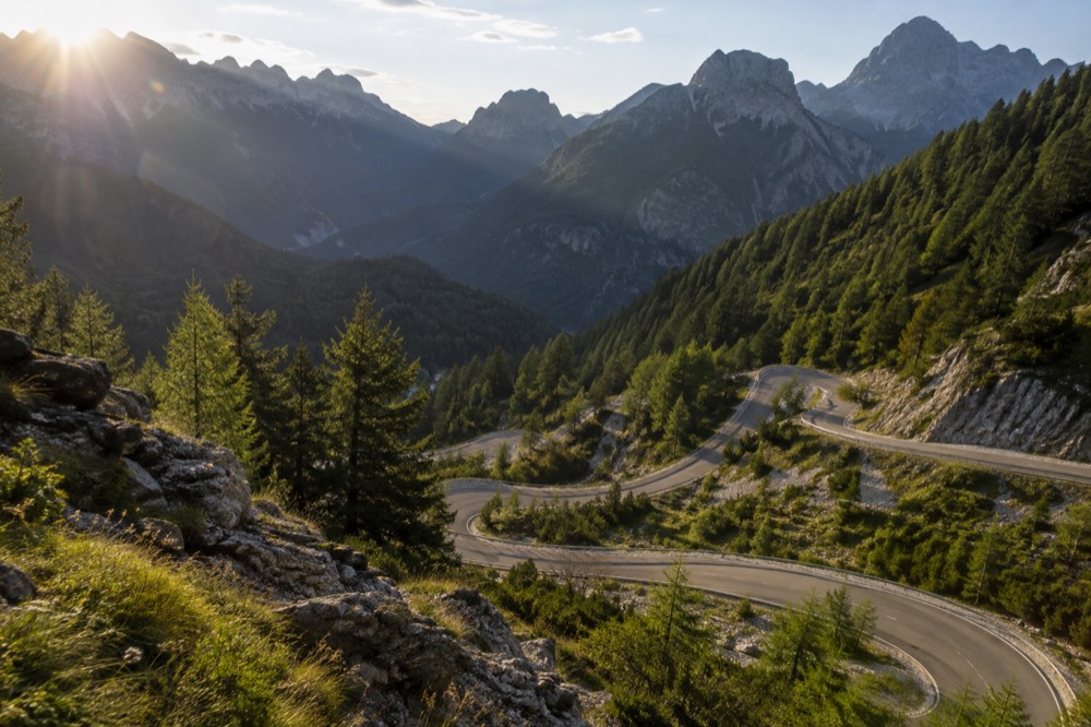 A steep alpine road with tight switchbacks cut into a green mountainside under a blue sky