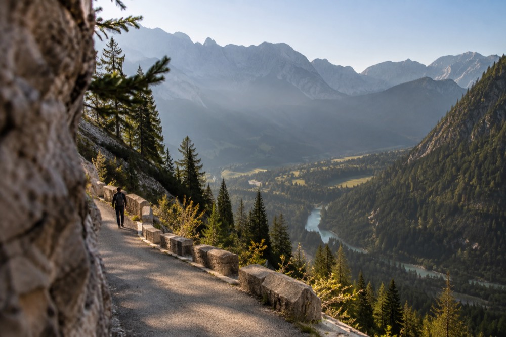 A steep road winding down a mountainside toward a turquoise river winding through a wide green valley