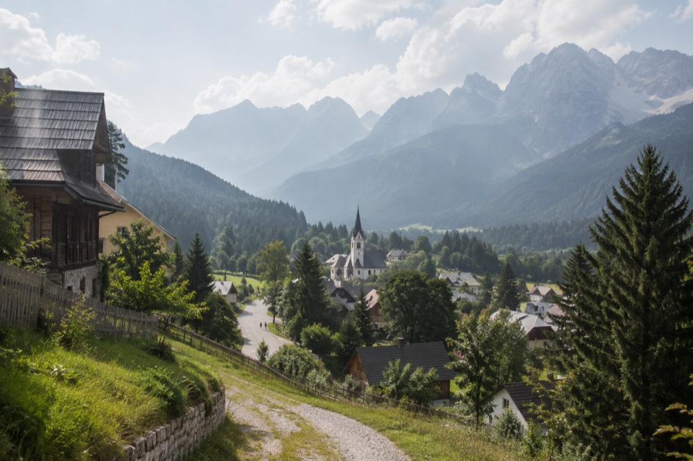 A small alpine town of wooden houses and a church spire in a green valley below high mountains